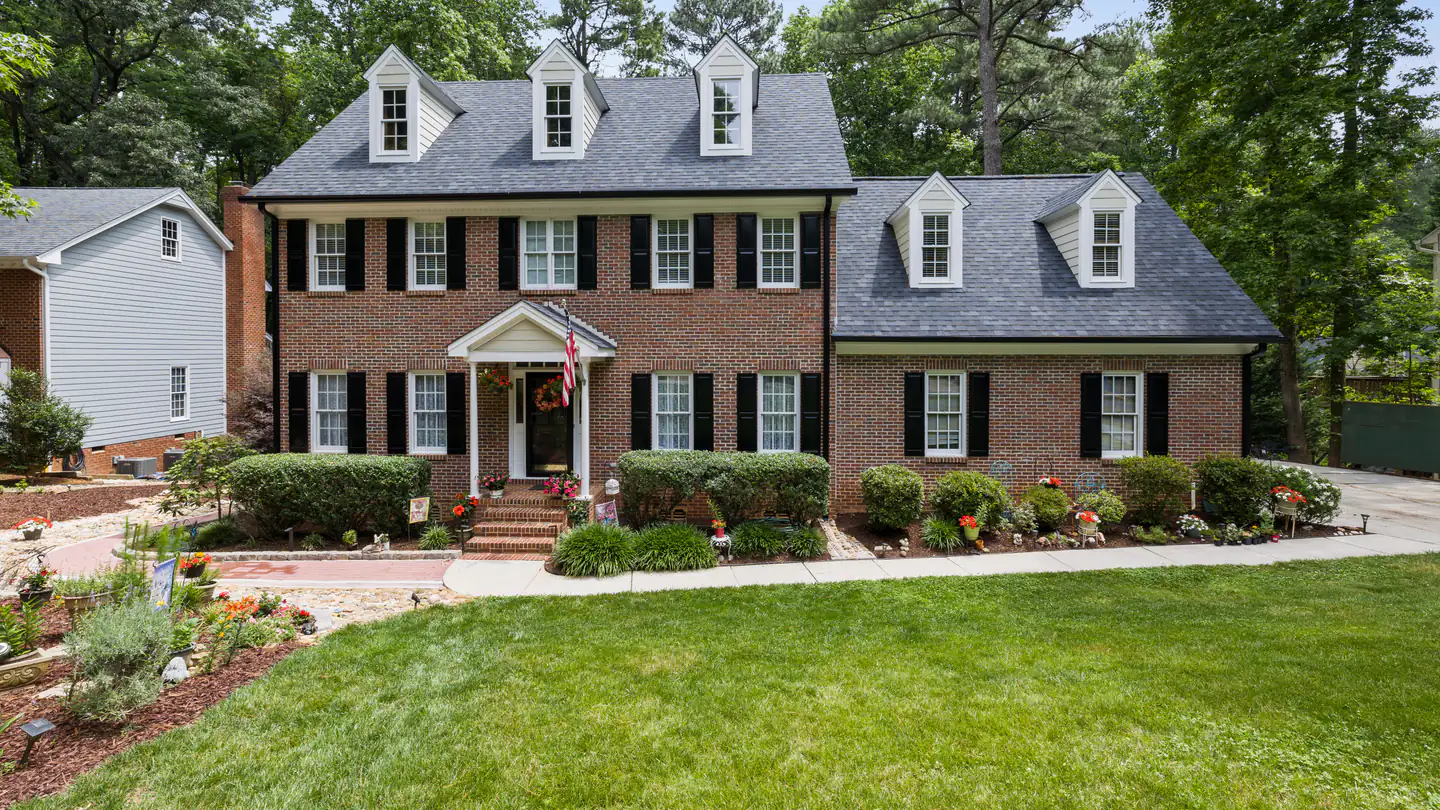 A red-brick colonial with dormer windows, black shutters and a clipped shrub border along the front.