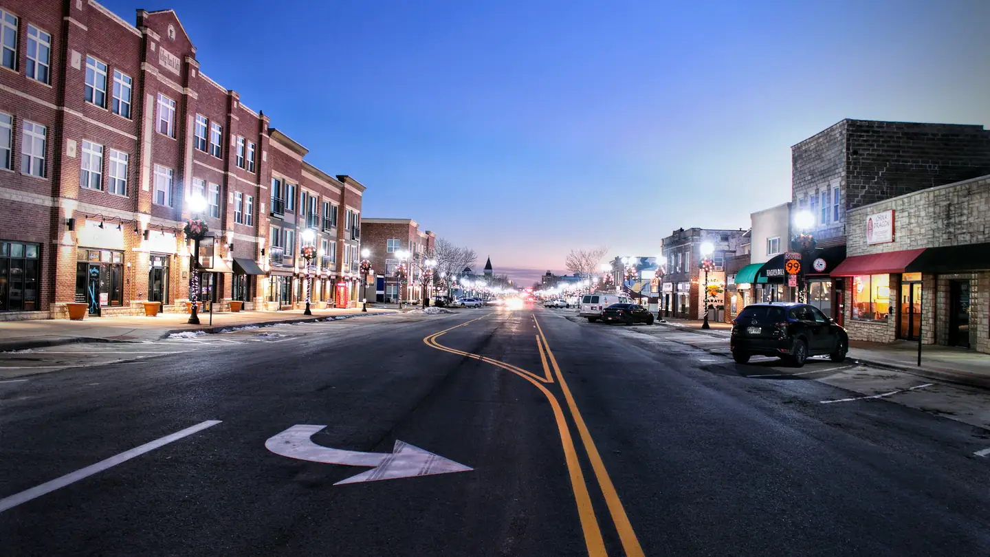 A small-town main street of brick storefronts at dusk, shop windows lit along both sides.