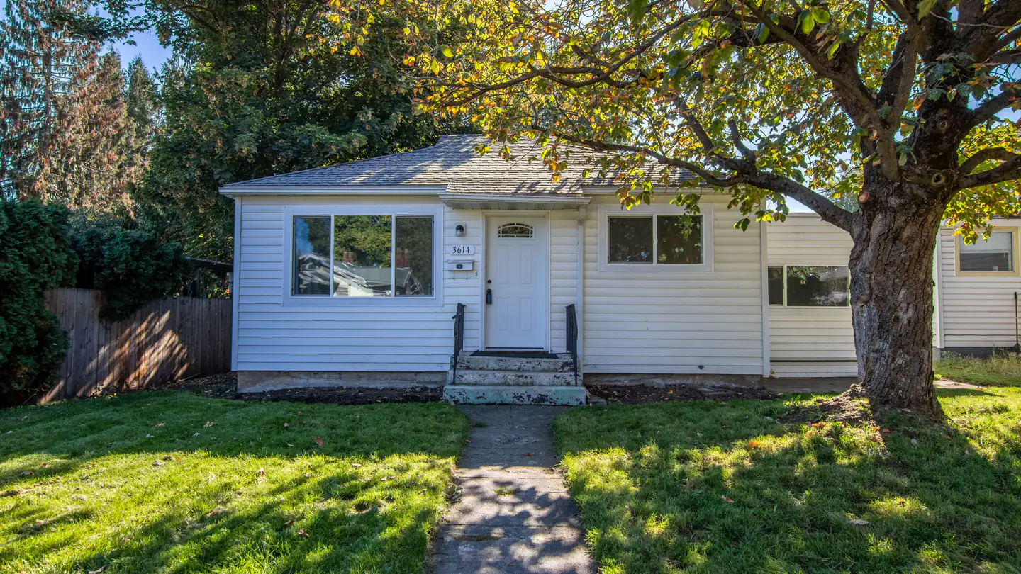 A single-story post-war ranch house with a picture window and a large shade tree in the front yard.