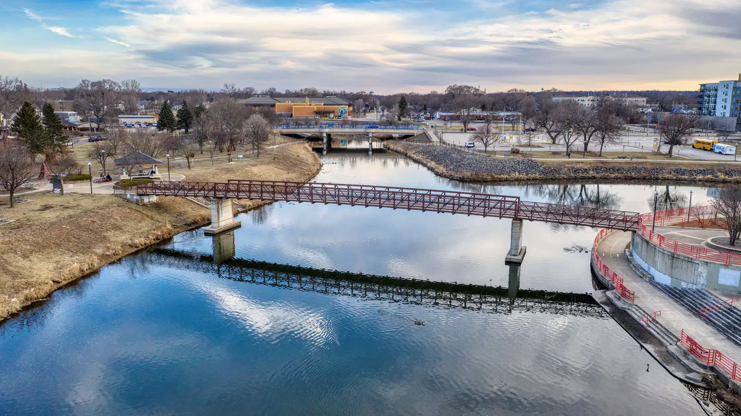A wooden pedestrian bridge crossing a calm river, with a small riverfront downtown behind it.
