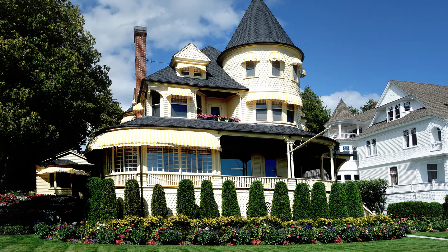 A yellow Queen Anne Victorian with a round corner turret and a wraparound porch, beside its white frame neighbor.